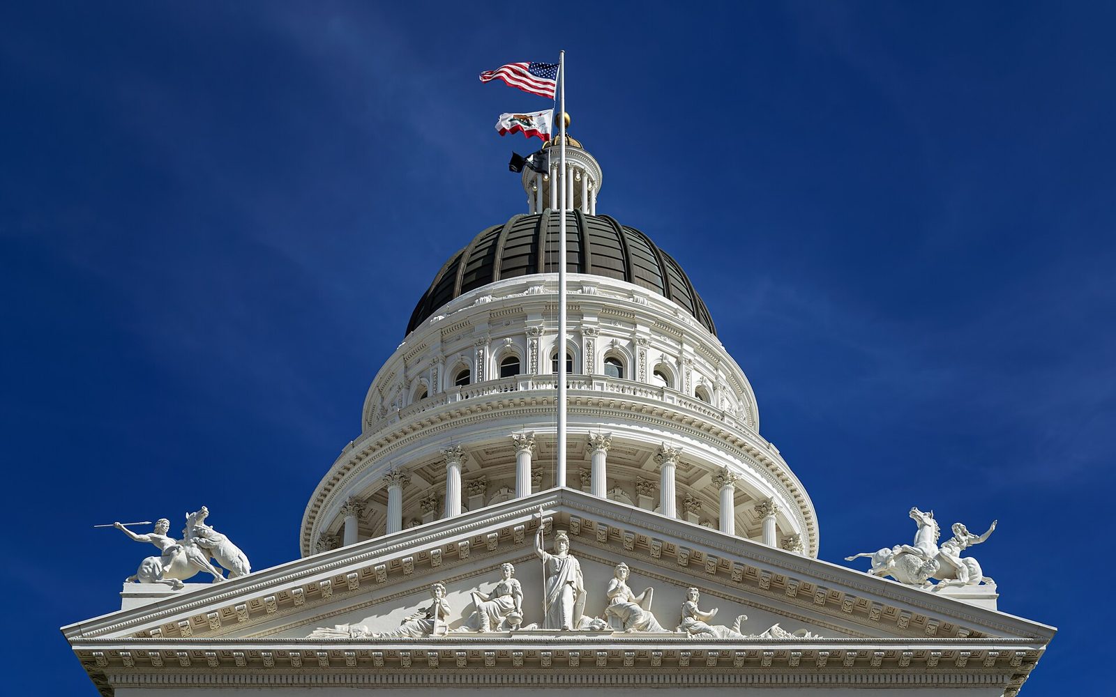 The dome of the California State Capitol in Sacramento against a deep blue sky, flags flying above the pediment.