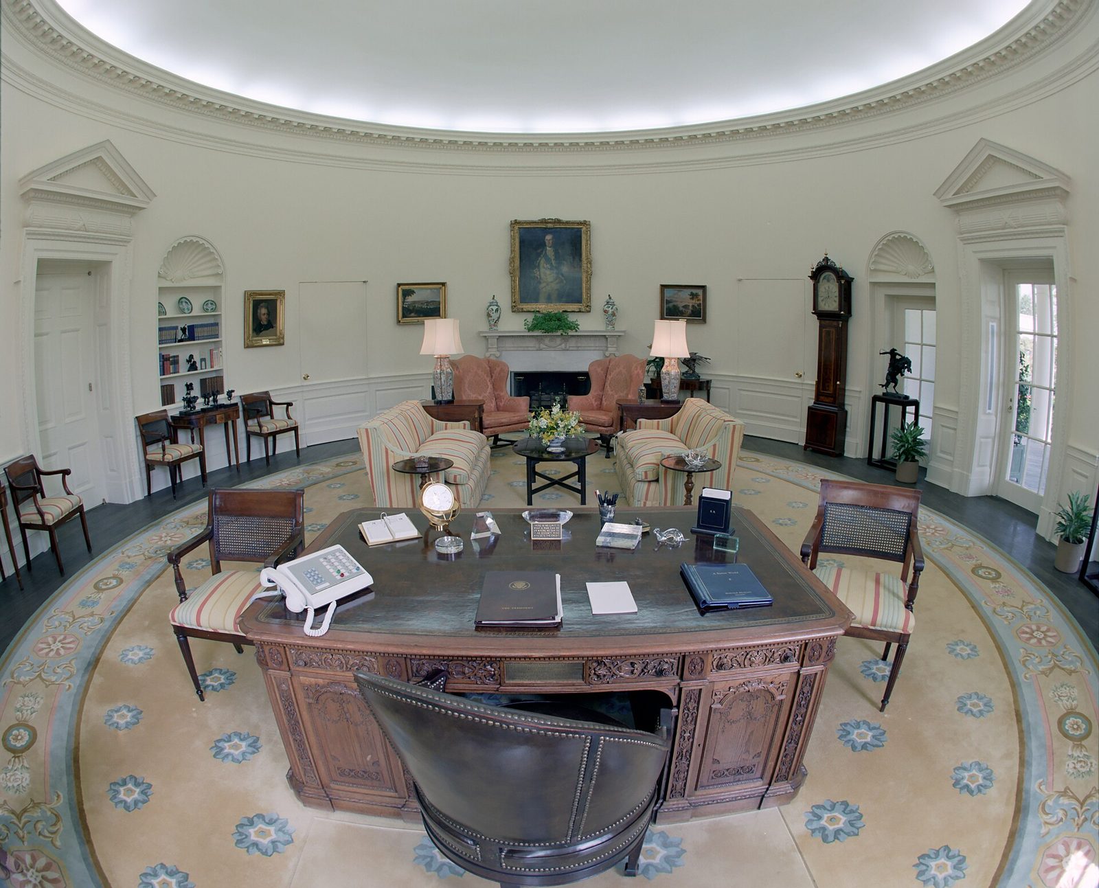 The Oval Office in the White House, empty: the Resolute desk, armchairs and a patterned rug under a curved white ceiling.