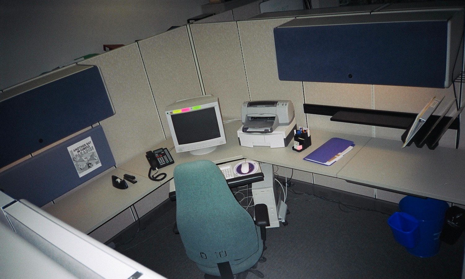 An empty office cubicle: a desk with an old computer monitor, a printer and a phone, and a chair pushed back from the desk.