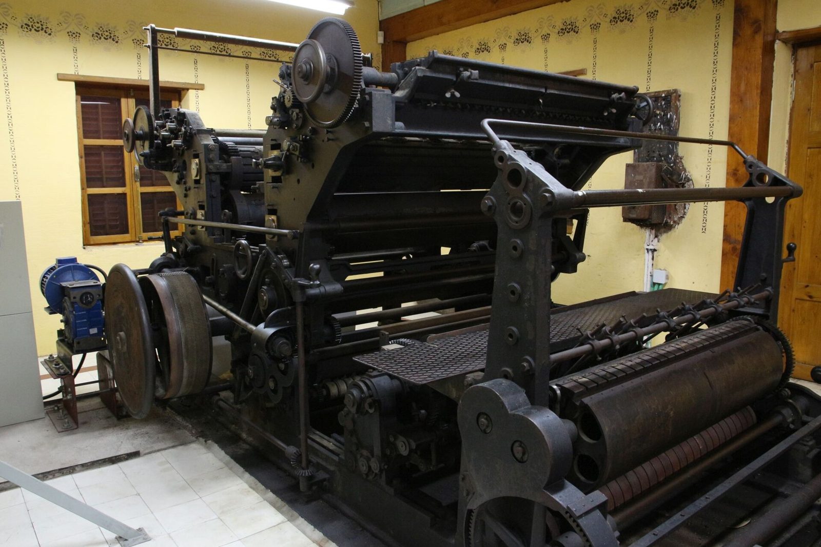 A large black cast-iron printing press with gears and rollers, preserved in a museum room with pale yellow walls.